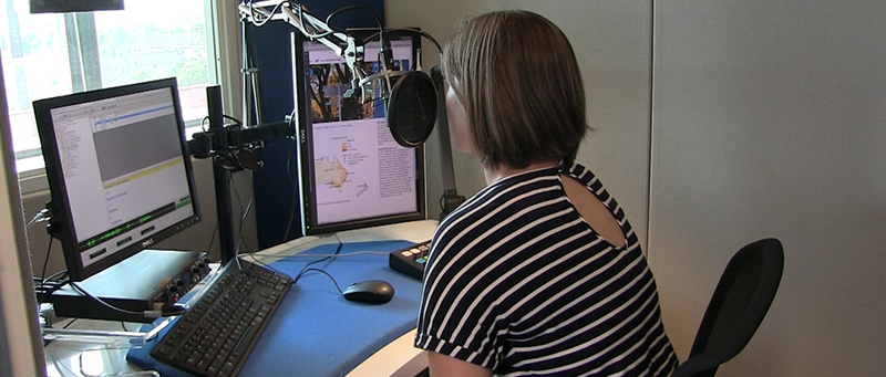 vrouw zit in een studio met haar rug naar de fotograaf en spreekt in een microfoon. Ze leest de tekst vanaf een beeldscherm.