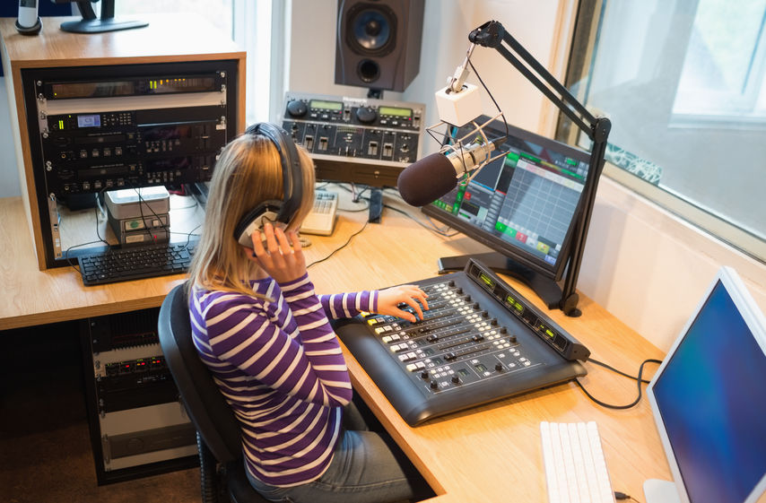 jonge vrouw in de studio met koptelefoon. Voor haar staat een mengpaneel, beeldscherm en microfoon.