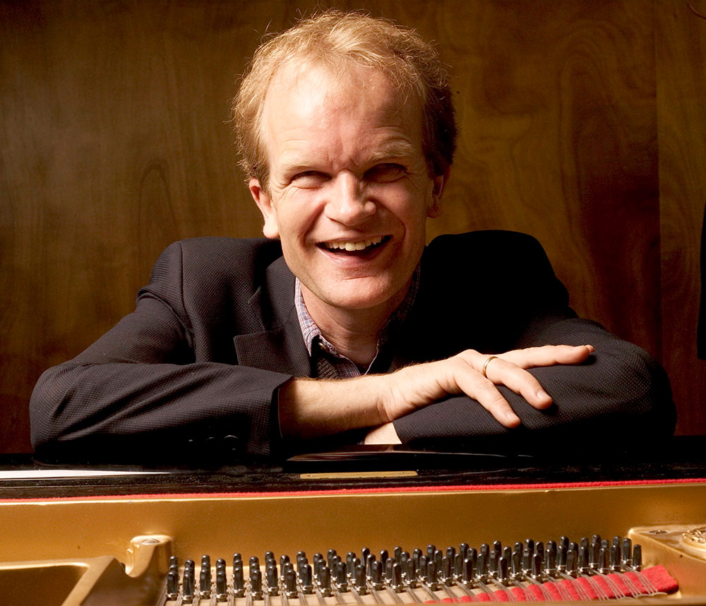 Bert van den Brink kijkt lachend in de camera en leunt op piano