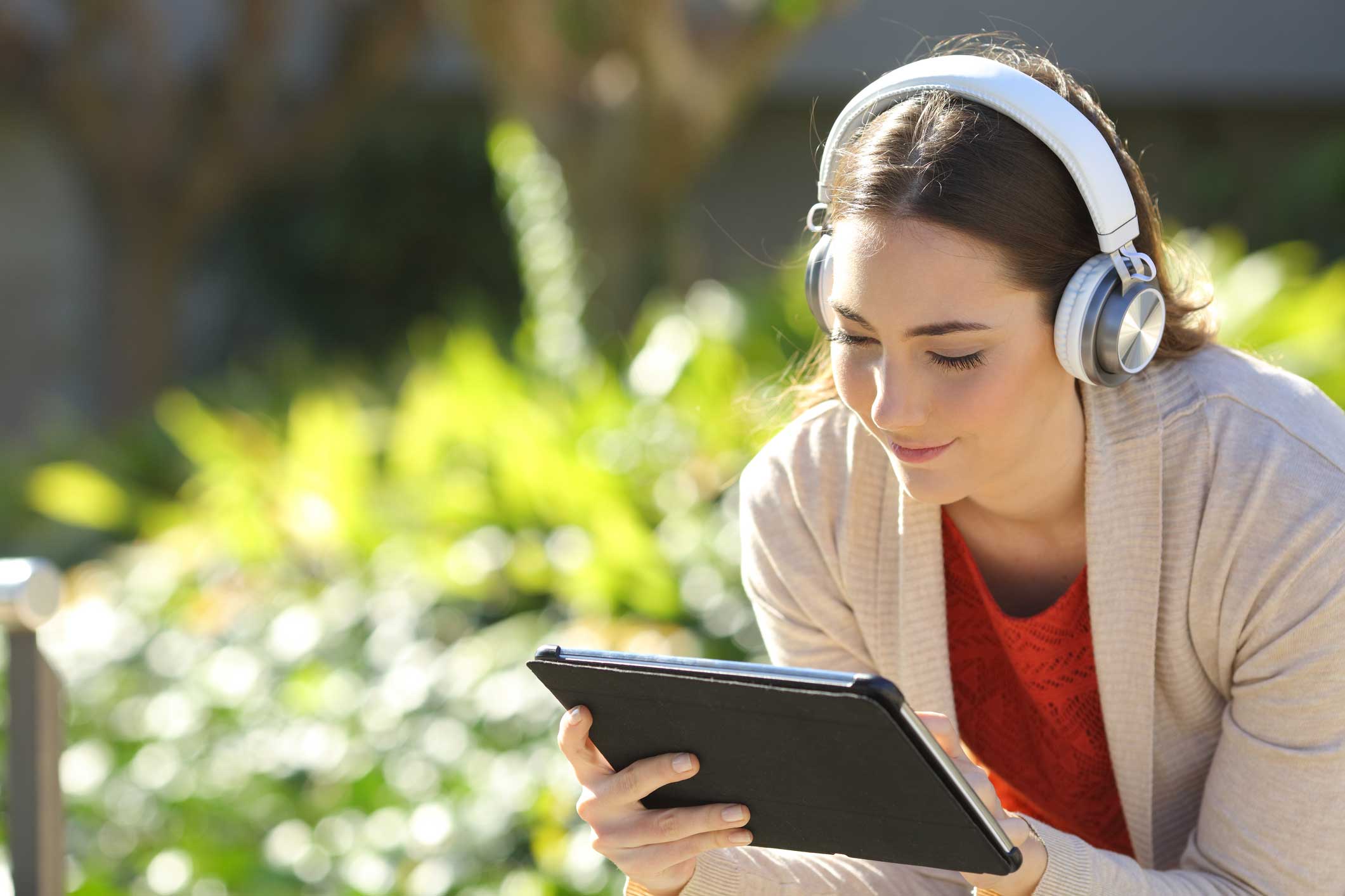 Een jonge vrouw luistert buiten met een koptelefoon op naar haar tablet.