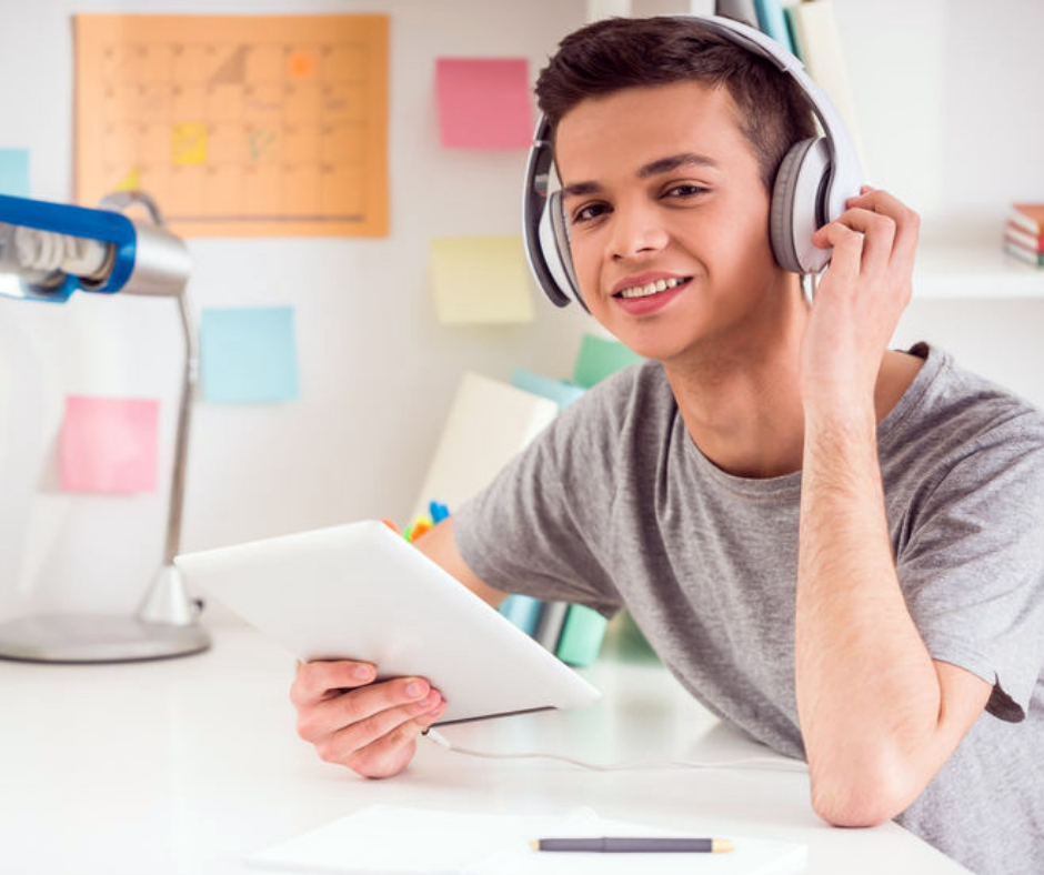 tiener jongen met koptelefoon zit aan bureau met laptop in een hand