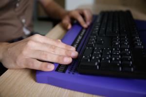 Twee handen tasten een brailleleesregel af. Op de brailleleesregel staat een toetsenbord