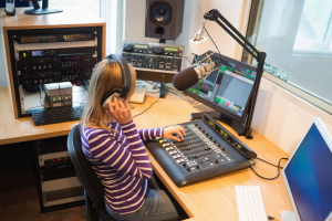 jonge vrouw in de studio met koptelefoon. Voor haar staat een mengpaneel, beeldscherm en microfoon.