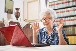 Een oudere dame zit voor een laptop en helft verheugd de handen omhoog.
