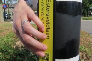 hand tastend op straatnaambordje in geel zwart contrast met braille, tekst en qr code, gemonteerd op verkeerslicht