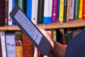 Een persoon houdt een e-reader vast; op de achtergrond een volle boekenkast.