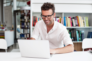 man werkt lachend met laptop. boekenkast op achtergrond