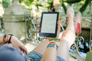 Een persoon zit buiten met de benen ophoog een e-boek op een e-reader te lezen.