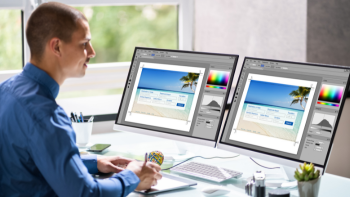 persoon werkt aan bureau met twee beeldschermen voor zich en daarop afbeelding van een tropisch oord